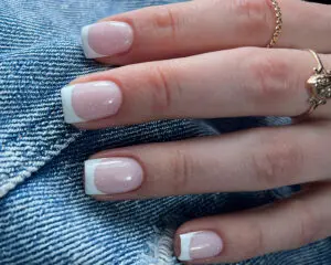 Manicure żelowy z białym klasycznym frenchem i brokatowym akcentem. Paznokcie pokryte naturalnym, przezroczystym żelem. Widoczny biały pasek przy końcu każdej płytki paznokcia, imitujący tradycyjny francuski manicure. Na paznokcie nałożono brokatowy top w kolorze holo, tworzący połyskujący akcent. Całość prezentuje się elegancko i nowocześnie.