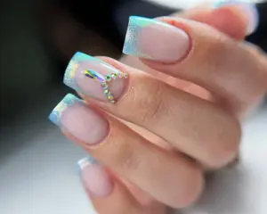 Manicure żelowy z jaskrawoniebieskim frenchem i pojedynczymi cyrkoniami. Paznokcie pokryte żelem w kolorze naturalnej płytki paznokcia. Na każdym paznokciu widać różowy pasek przy końcu płytki - francuski kolorowy akcent. Na niektórych paznokciach umieszczono po jednym cyrkonie, dodającym elegancji.