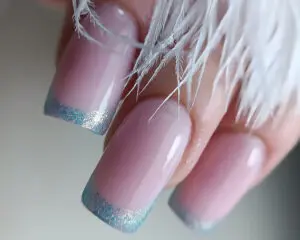Zdjęcie przedstawia dłonie eleganckiej kobiety z manicure w stylu klasycznego frencha, lecz zamiast typowych różowych pasków przy górnej granicy płytki paznokciowej widnieją brokatowe zakończenia. Paznokcie są długie, w kształcie kwadratu pokryte różowym lakierem. Na różowym tle błyszczą brokatowe drobinki brokatu tworzące cienkie paski przy samych końcówkach paznokci. Ten ozdobny manicure dodaje typowemu frenchowi stylu i oryginalności. Podkreśla elegancki wygląd zadbanej dłoni.