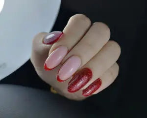 Zdjęcie przedstawia dłonie młodej kobiety z manicure w stylu french, lecz zamiast typowego różowego paska u góry płytki paznokcia, ma ona czerwone zakończenia. Paznokcie są średniej długości, owalnego kształtu. Ten manicure dodaje typowemu frenchowi nutkę wyrazistości i nowoczesności. Podkreśla zadbaną dłoń.
