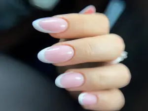 Zdjęcie przedstawia dłonie kobiety z pomalowanymi paznokciami w stylu klasycznego french manicure'u. Paznokcie są wydłużone i mają kształt migdałowy. Na białym tle płytki paznokciowej znajduje się różowy pasek przebiegający u góry, imitujący naturalną linię u nasady płytki.