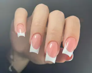 Zdjęcie przedstawia dłoń kobiety z długimi, wydłużonymi paznokciami w stylizacji French manicure. Paznokcie są pomalowane na jasny kolor z wyrazistą, białą końcówką imitującą naturalny uśmiech paznokcia. Jest to klasyczny, elegancki manicure.