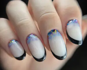 Zdjęcie przedstawia zadbaną dłoń kobiety z owalnymi paznokciami pomalowanymi w stylu french manicure. Paznokcie pokrywa biały lakier z wyjątkiem czarnych, błyszczących pasków biegnących przy górnych krawędziach płytek. Kontrast czerni i bieli dodaje temu manicure nowoczesnego i nieco rockowego charakteru. Owalny kształt podkreśla kobiecy, elegancki wygląd dłoni. Czarna linia imituje naturalny wygląd płytki paznokciowej, podkreślając zadbane paznokcie.